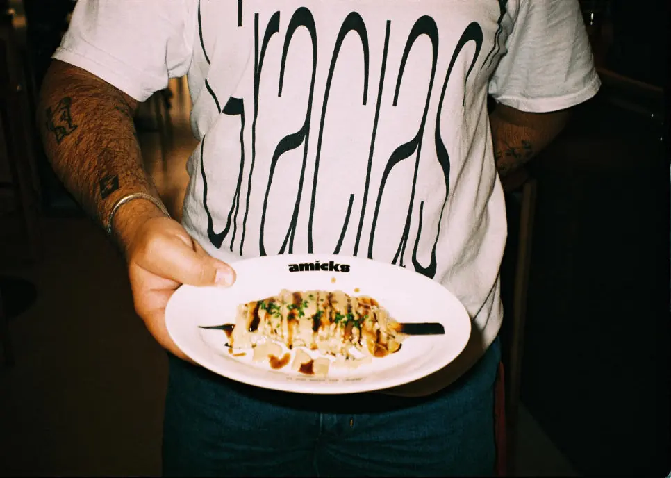 Un cambrer amb una samarreta que diu Gracias sosté una broqueta de céleri en un plat amb el logo del Bar Amicks