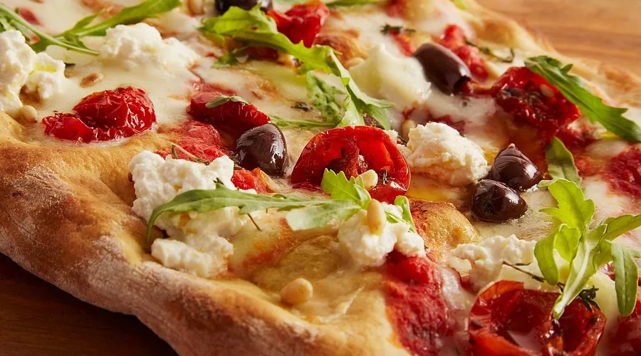 Pizza con tomates asados, aceitunas negras, ricotta, piñones y rúcula, de cerca