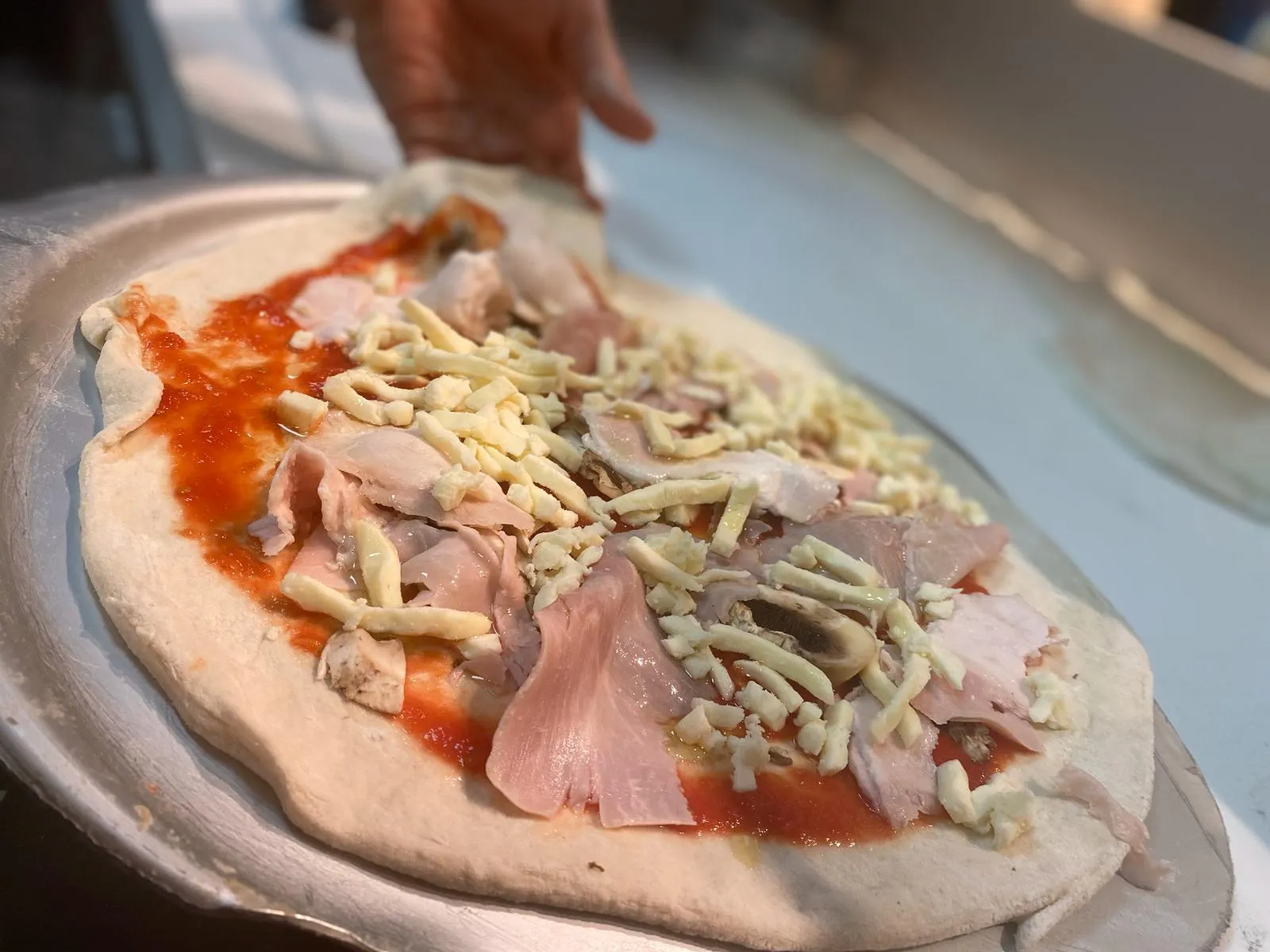 Una pizza se prepara a mano sobre la pala: la masa estirada, con tomate, jamón y queso