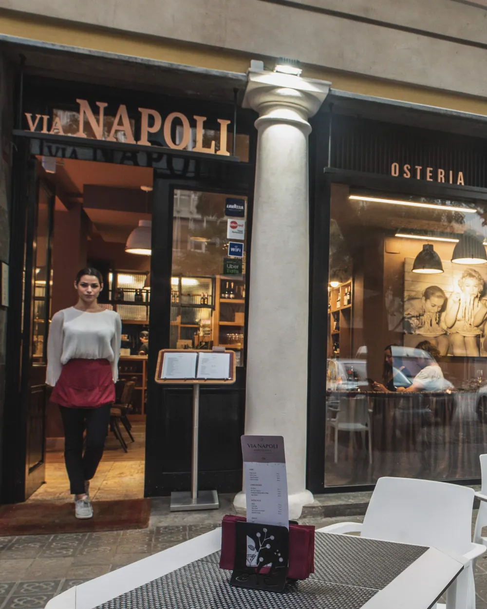 La fachada de Via Napoli en la calle Nàpols, con la terraza delante y una camarera en la puerta